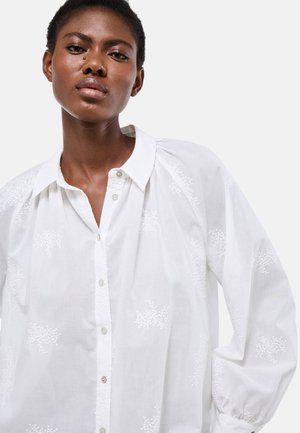 Femme portant un chemisier blanc à boutons avec une broderie florale subtile et des manches bouffantes, posant devant un fond clair uni.