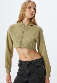 Sudadera cortada verde oliva con bordado de mariposa en la parte frontal, cierre de cremallera y mangas largas, combinada con jeans negros de cintura alta.