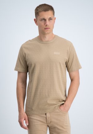 Man met kort haar die een beige T-shirt met patroon en beige broek draagt, staand met één hand in de zak tegen een effen achtergrond.