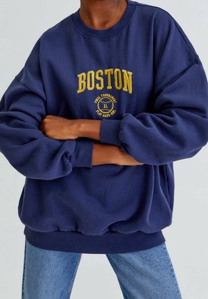 Osoba ubrana w luźną granatową bluzę z napisem "BOSTON" i okrągłym emblematem w żółtych literach, w połączeniu z niebieskimi dżinsami. Ręce skrzyżowane.