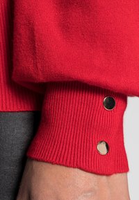 Roter Strickpulloverärmel mit geripptem Muster, goldenen Knopfakzenten nahe dem Bündchen und glattem Stoff, der bis zum Handgelenk reicht.