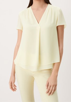 Frau trägt eine hellgelbe kurzärmelige V-Ausschnitt-Bluse mit einer Frontfalte, kombiniert mit hellgelben Hosen vor einem schlichten Hintergrund.