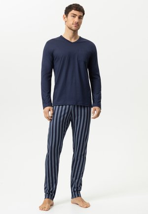 Mann steht barfuß, trägt ein marineblaues Langarmshirt und blaue gestreifte Pyjamahosen vor weißem Hintergrund.