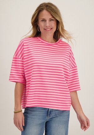 Vrouw die lacht, draagt een losse roze en lichtroze gestreepte t-shirt met ellebooglange mouwen en blauwe spijkerbroek tegen een effen achtergrond.