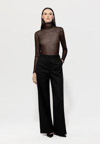 Braun-transparentes Rollkragenoberteil mit subtilen Verzierungen, kombiniert mit hochtaillierten schwarzen weit geschnittenen Hosen. Das Model steht mit einer Hand in der Tasche.