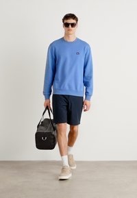 Light blue sweatshirt, navy shorts, beige sneakers. Model carries a black duffel bag with silver hardware. Smooth textures, casual design.