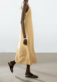 Gelbes Leinenärmelloses Kleid mit lockerer Passform und seitlichen Schlitzen, kombiniert mit flachen schwarzen Sandalen. Glatte Textur, schlichtes Design.