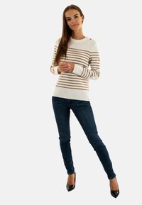 Pull blanc avec des rayures beiges, col rond et boutons aux épaules ; associé à un jean bleu foncé et des escarpins noirs à bout pointu.