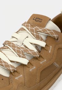 Καφέ σουέντ αθλητικό παπούτσι με εκρού λεπτομέρειες. Διαθέτει υφασμάτινη υφή, λουριά σε σχήμα σταυρού και λογότυπο στη γλώσσα. Καλαίσθητες ραφές.