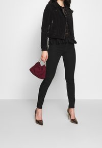 Donna che indossa una giacca in suede nera, un top in pizzo nero, pantaloni skinny neri e tacchi alti con stampa serpente, mentre tiene in mano una piccola borsa bordeaux.