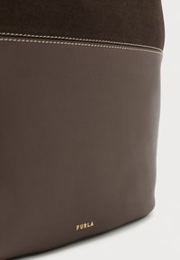 Sac en cuir marron avec une partie supérieure en tissu texturé marron foncé. Présente des coutures blanches contrastantes et un logo doré en relief à la base.