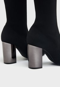 Gros plan sur des bottines chaussettes noires avec des talons blocs métalliques brillants sur un fond clair.