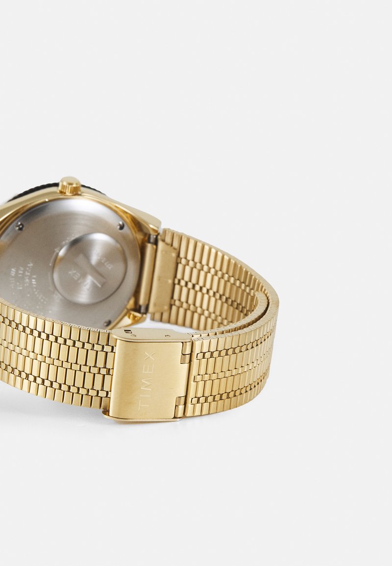 Reloj de pulsera de metal dorado con una correa texturizada, que presenta una caja redonda y un fondo metálico liso. Cierre marcado como "TIMEX" para un cierre seguro.