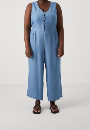 Femme portant une combinaison en denim bleu clair sans manches avec des boutons sur le devant, associée à des sandales noires à brides, debout sur un fond uni.