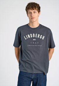 Tummanharmaa puuvillapaita, jossa on valkoiset tekstit "LINDBERGH" vuoristoaiheen kanssa ja "1927 KOPENHAGENA" sen alla. Lyhyet hihat.