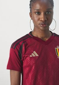 Sarkans futbola krekls ar melniem akcentiem, zelta Adidas logo, dimanta rakstu un nacionālo emblēmu uz krūtīm. Īsas piedurknes un V veida kakla izgriezums.