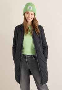 Pikowana czarna kurtka z zamkiem błyskawicznym, noszona na jasnosprawnym swetrze. Model ma na sobie również zieloną prążkowaną czapkę z metką. Szare dżinsy.
