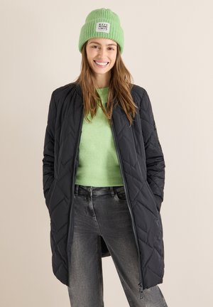 Quiltad svart jacka med dragkedja, bärs över en ljusgrön tröja. Modellen har även på sig en ribbad grön mössa med en etikett. Grå jeans.