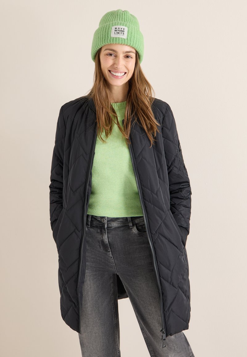 Pikowana czarna kurtka z zamkiem błyskawicznym, noszona na jasnosprawnym swetrze. Model ma na sobie również zieloną prążkowaną czapkę z metką. Szare dżinsy.