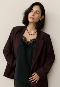 Burgundowy żakiet na czarnym topie z koronkowym wykończeniem, z naszyjnikiem z czerwonym kamieniem. Stylizacja charakteryzuje się gładkimi teksturami i warstwowymi wzorami.