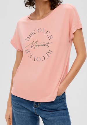 Kobieta w różowej koszulce z podwiniętymi rękawami i srebrnym napisem „Discover Recover, Enjoy the Moment”, w połączeniu z niebieskimi dżinsami.
