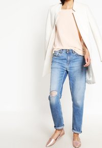 Kvinna klädd i ljusblå slitna jeans, ljusrosa topp, vit öppen kavaj och rosa spetsiga ballerinas, stående mot en vit bakgrund.