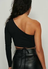 Schwarzes Crop-Top mit einem langen Ärmel und einer freien Schulter, das eine figurbetonte Passform aufweist und an der Taille mit einem Knoten versehen ist. Hergestellt aus einem glatten Stoff.