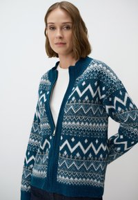 Gilet en maille teal avec des motifs géométriques blancs et gris. Présente une fermeture éclair à l'avant et un col côtelé, fabriqué en mélange de laine texturé.