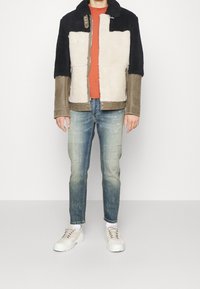 Braun, Schwarz und cremefarbene, color-blocked Shearlingjacke mit einem Reißverschluss vorne, Metallbeschlägen und einer plüschigen Textur; kombiniert mit hellblauen, verwaschenen Jeans und weißen Sneakern.