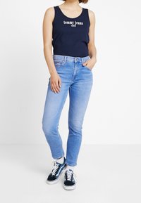 Granatowa koszulka bez rękawów z logo "Tommy Jeans", zestawiona z jasnoniebieskimi dżinsami i czarnymi sneakersami z białymi sznurówkami i niebieskimi akcentami.