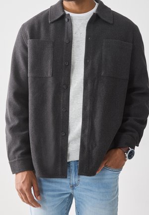 Mężczyzna noszący ciemnoszarą koszulową kurtkę z guzikami na jasnoszarej bluzie i jasnoniebieskich dżinsach, z zegarkiem na lewym nadgarstku i ręką w kieszeni.