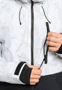 Weiß und grau gemusterte Jacke mit Kapuze, ausgestattet mit einem schwarzen Reißverschluss, schwarzen Bündchen und einer Seitentasche mit verstellbarem Kordelzug.