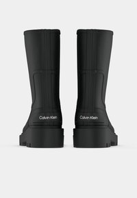 Črne gumijaste škornje do sredine meč z debelimi podplati, zadnji pogled, s "Calvin Klein" logotipom na petah na belem ozadju.