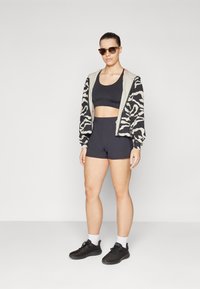 Kraftig sort hættejakke med et beige og sort zebra mønster, sort sportstop, højtaljede sorte shorts og sorte træningssko med hvide sokker.