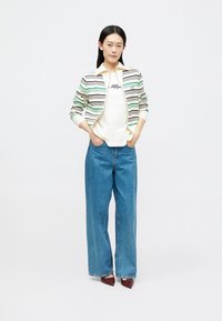 Jachetă din bumbac cu fermoar, cu dungi verzi și negre, purtată peste un tricou alb, asortată cu blugi largi din denim albastru și pantofi cu toc bordeaux.