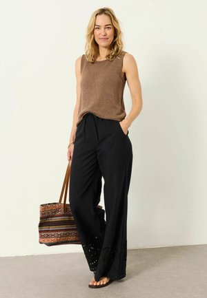 Frau mit blonden Haaren, die ein ärmelloses braunes Oberteil und weite schwarze Hosen trägt, hält eine gemusterte Tragetasche und steht vor einer weißen Wand.