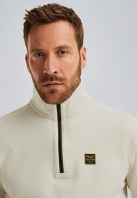 Sudadera de medio cierre en color beige claro con un alto cuello, que presenta una cremallera negra y un pequeño parche de logo negro y dorado en el pecho.