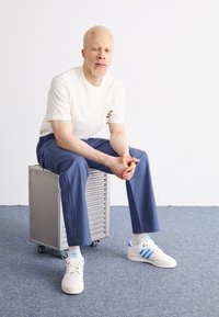 Vit t-shirt med en liten färgglad logotyp, blå byxor och vita sneakers med blå ränder. Sitter på en metallbox.