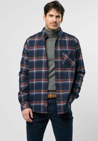 Donkerblauw geruit flanellen overhemd met rode en witte strepen, met een puntige kraag en borstzak, gelaagd over een grijze turtleneck.