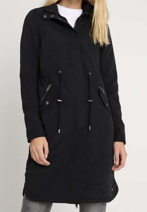 Femme portant un long manteau noir zippé avec cordons à la taille et poches avant zippées, par-dessus une chemise blanche et un pantalon gris.