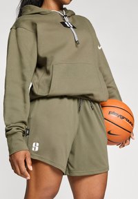 Haut à capuche vert olive avec cordon et poche avant, associé à un short assorti avec un design texturé et un logo imprimé. Tient un ballon de basket.