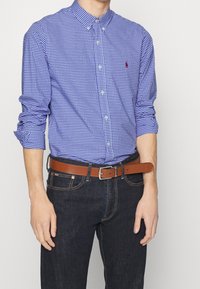 Niebieska i biała koszula w kratkę z guzikami, z długimi rękawami podwiniętymi, w połączeniu z ciemnoniebieskimi dżinsami i brązowym skórzanym paskiem. Obecny detal logo w kolorze czerwonym.