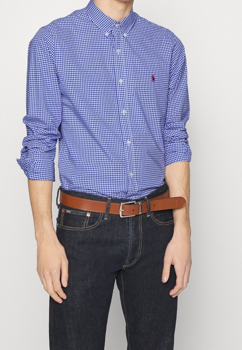 Niebieska i biała koszula w kratkę z guzikami, z długimi rękawami podwiniętymi, w połączeniu z ciemnoniebieskimi dżinsami i brązowym skórzanym paskiem. Obecny detal logo w kolorze czerwonym.