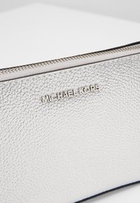 Pochette en cuir argenté texturé avec une fermeture éclair sur le dessus et un logo métallique Michael Kors au centre avant.