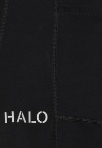 Svarta tygbyxor med synlig sömnad och "HALO"-logotyp i vitt. Inkluderar fickor och har en jämn textur. Närbild.