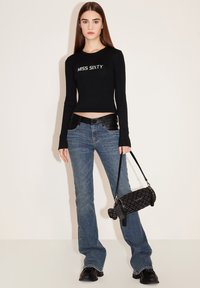 Schwarzes geripptes Crop-Top mit "MISS SIXTY"-Aufschrift, dunkelblaue Schlagjeans und eine schwarze gesteppte Handtasche. Model trägt schwarze Schuhe.
