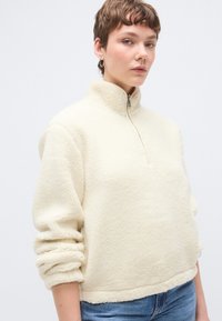 Pulover od krem flisa s visokim ovratnikom, zatvaračem na patentni zatvarač i manžetama na rukavima. Mekana tekstura s opuštenim krojem, uparena s trapericama.