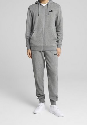 Person iført en grå Puma træningsdragt med lynlåshætte og joggingbukser, parret med hvide sneakers, stående på ensfarvet baggrund.