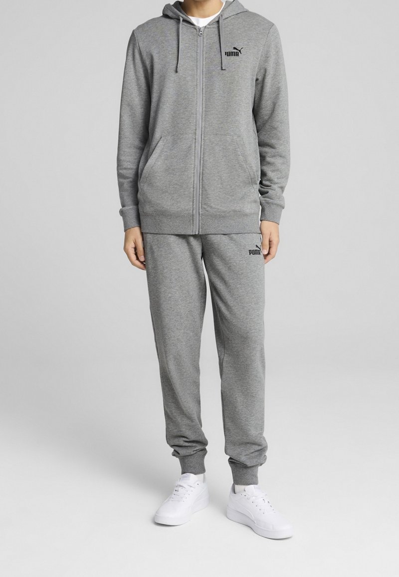 Personne portant un survêtement gris Puma avec un sweat à capuche zippé et un pantalon de jogging, assorti à des baskets blanches, debout sur un fond uni.