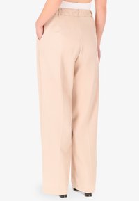 Pantaloni a gamba larga color beige chiaro con una texture liscia, dotati di vita alta e tasche laterali. Cuciture visibili e dettagli sartoriali.